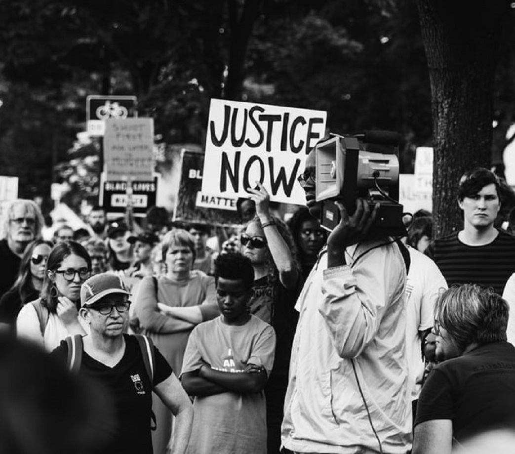 black and white image of a protest, with a sign reading 'Justice Now'