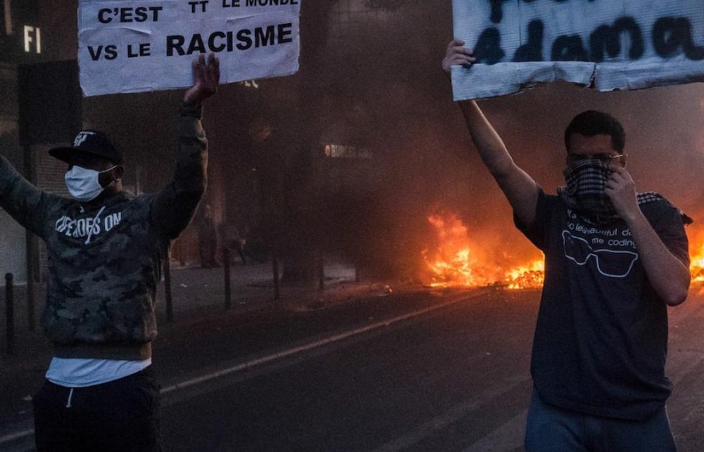Two people in masks hold up signs in front of a fire.