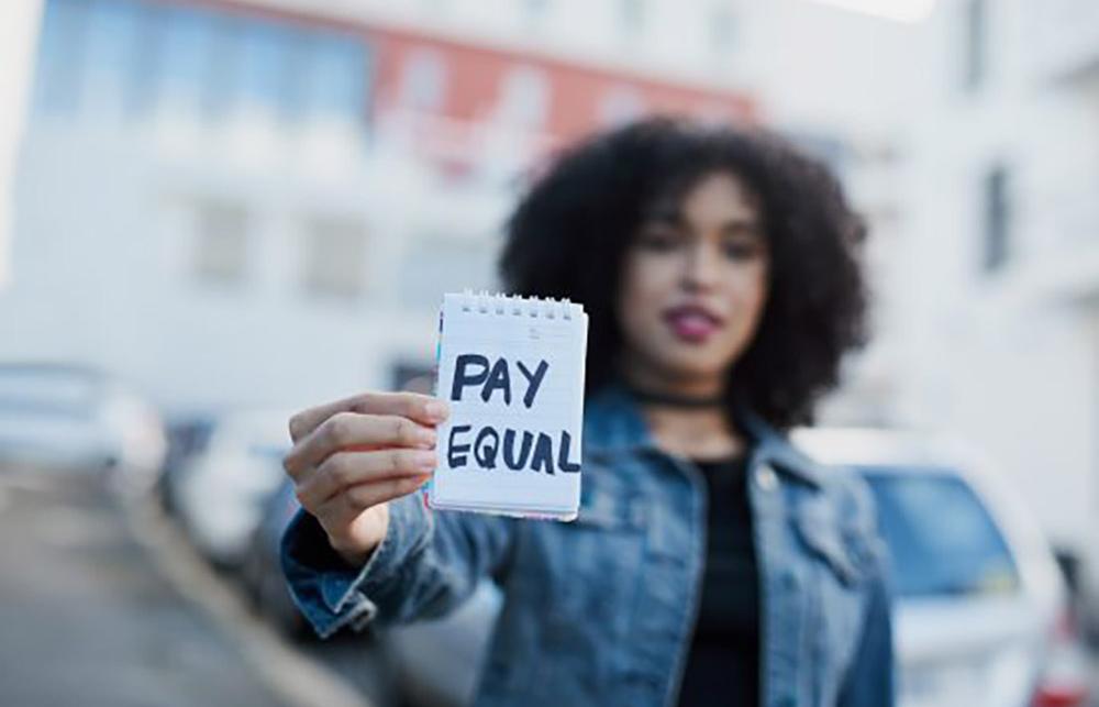 Someone holds a piece of paper reading 'Pay Equal'